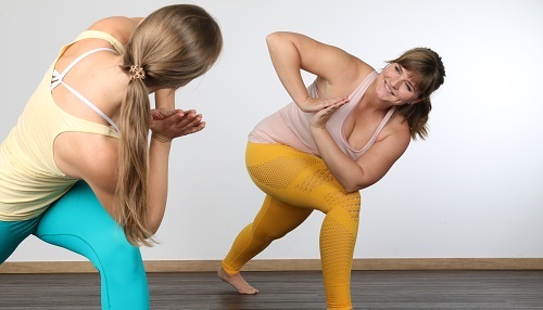 Yoga Gutschein verschenken heißt Freude verbreiten: Zwei Frauen lächeln sich an im gedrehten High Lunge