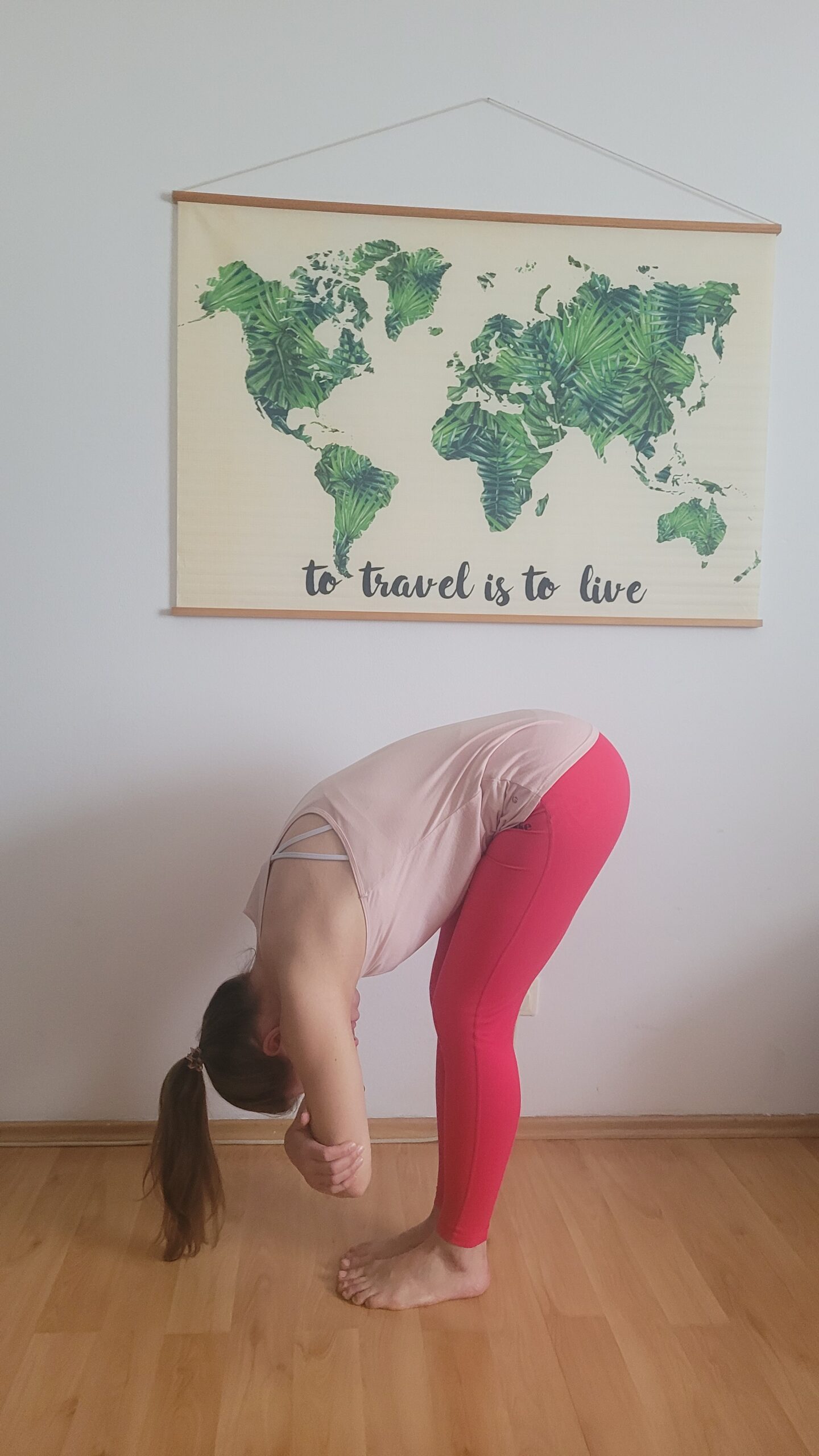 Yogalehrerin in stehender Vorbeuge mit verschränkten Armen vor Wandbild mit Weltkarte im Blattmuster