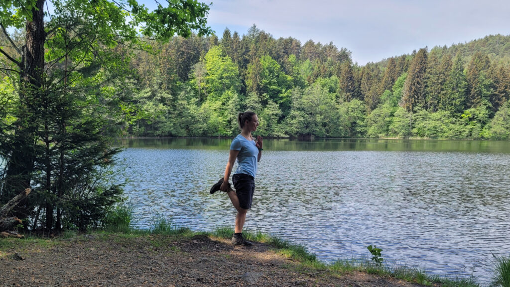 Frau steht auf einem Bein und dehnt Oberschenkel am See im Wald