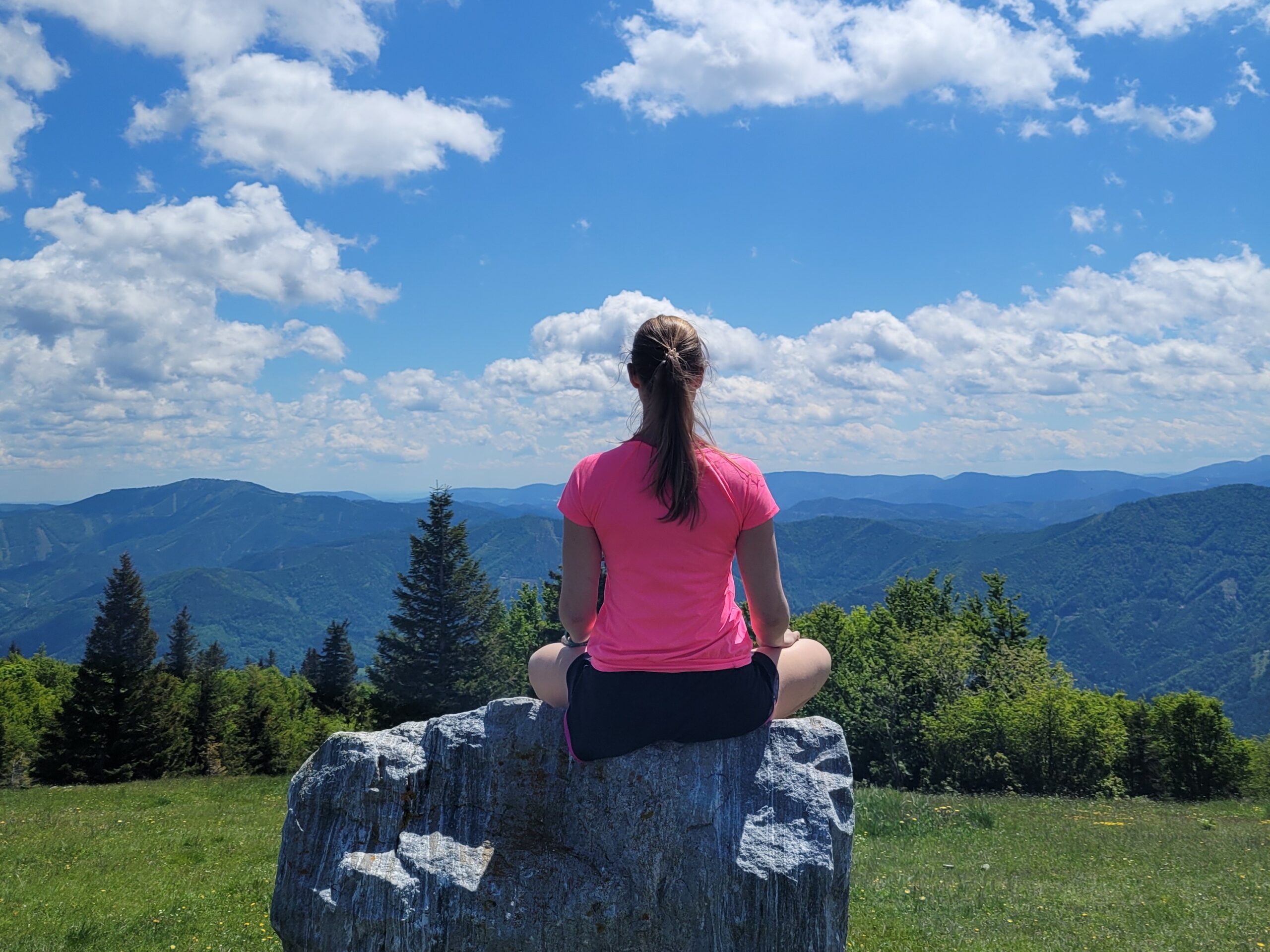 Frau von hinten in sommerlichem Sportoutfit im Meditationssitz auf einem Felsen in den Bergen integriert 5 Minuten Achtsamkeit in ihre Wanderung