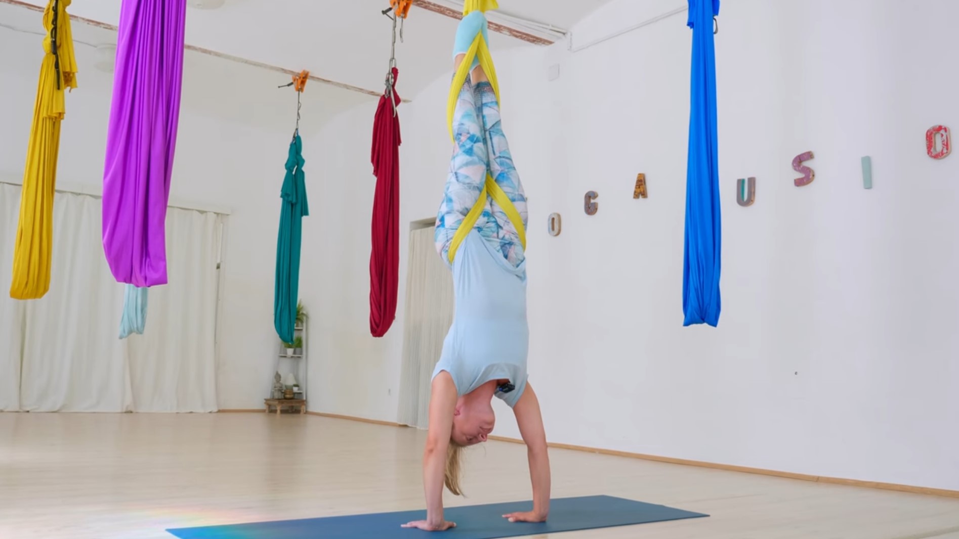 Aerial Yoga Lehrerin im Handstand mit einem der bunten Tücher, an der Wand der Yogafusion Schriftzug