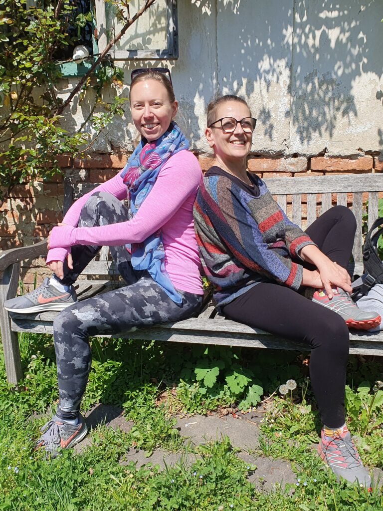 Fastenleiterin Angelika Hager und Yogalehrerin Sabrina Farkas sitzen lächelnd Rücken an Rücken auf einer Gartenbank