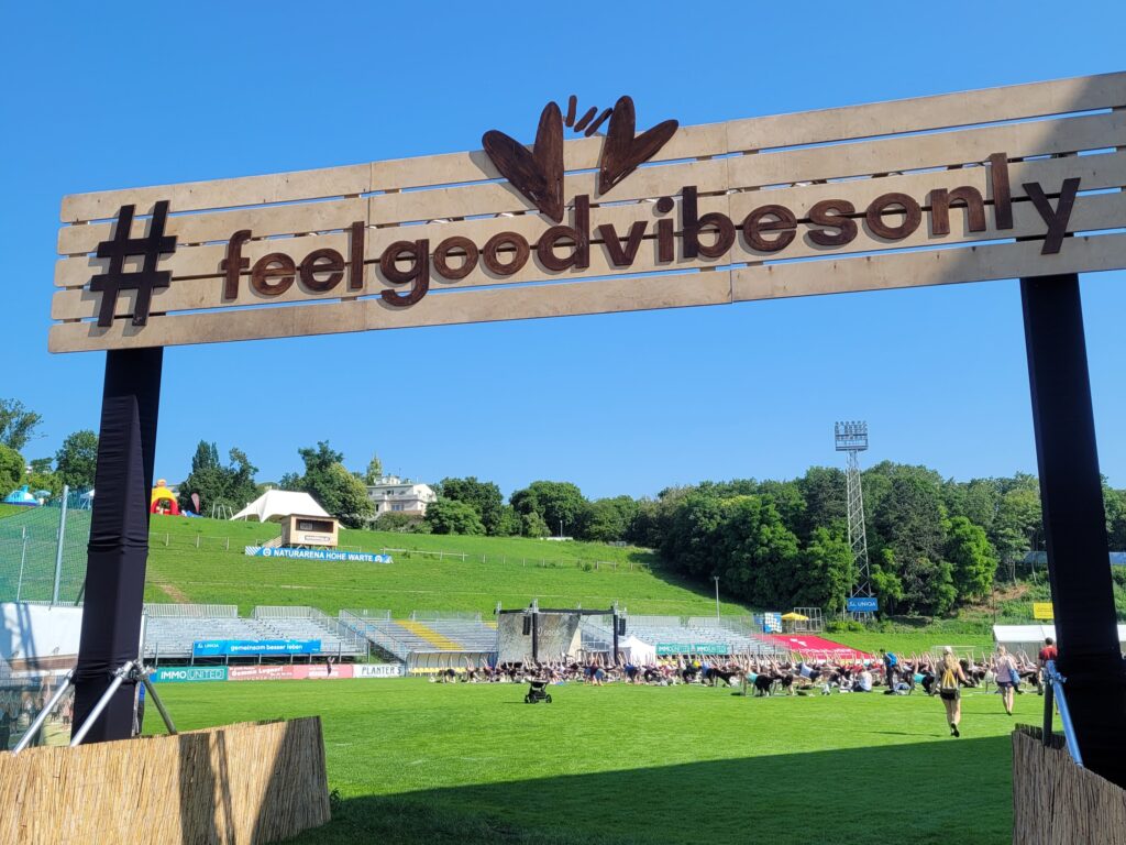 Eingangsbereich des Feel Good Festival mit #feelgoodvibesonly Schild im Vordergrund und Wiese mit Bühne und Yogis im Hintergrund