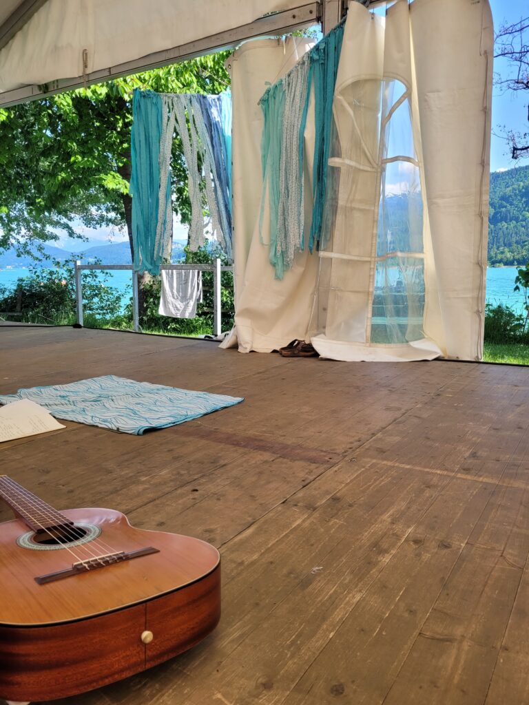 Gitarre auf Holzboden in weißem Yoga Festival Zelt, dahinter grüne Natur und türkisblauer See