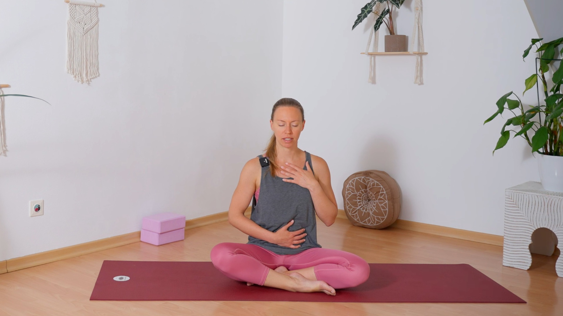 Frau in Yogakleidung sitzt mit gekreuzten Beinen auf Yogamatte, die rechte Hand auf dem Bauch und die linke am Herzen, während sie mit geschlossenen Augen die volle Yoga Atmung übt
