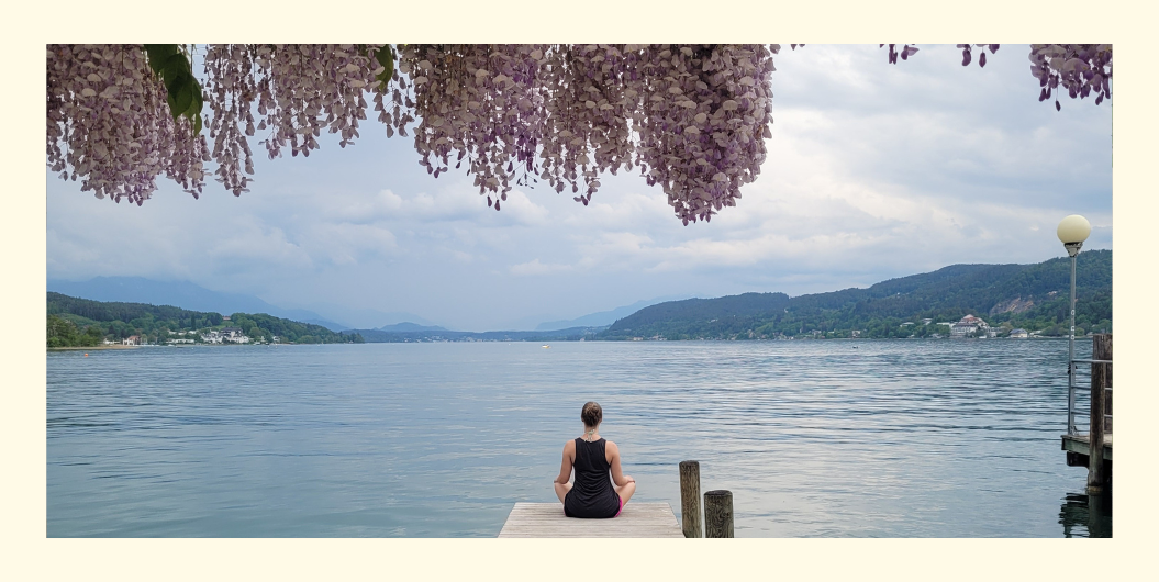 Rückenansicht einer Frau bei einer geführten Meditation im Sitzen am Steg eines Sees, über ihr Glyzinienblüten