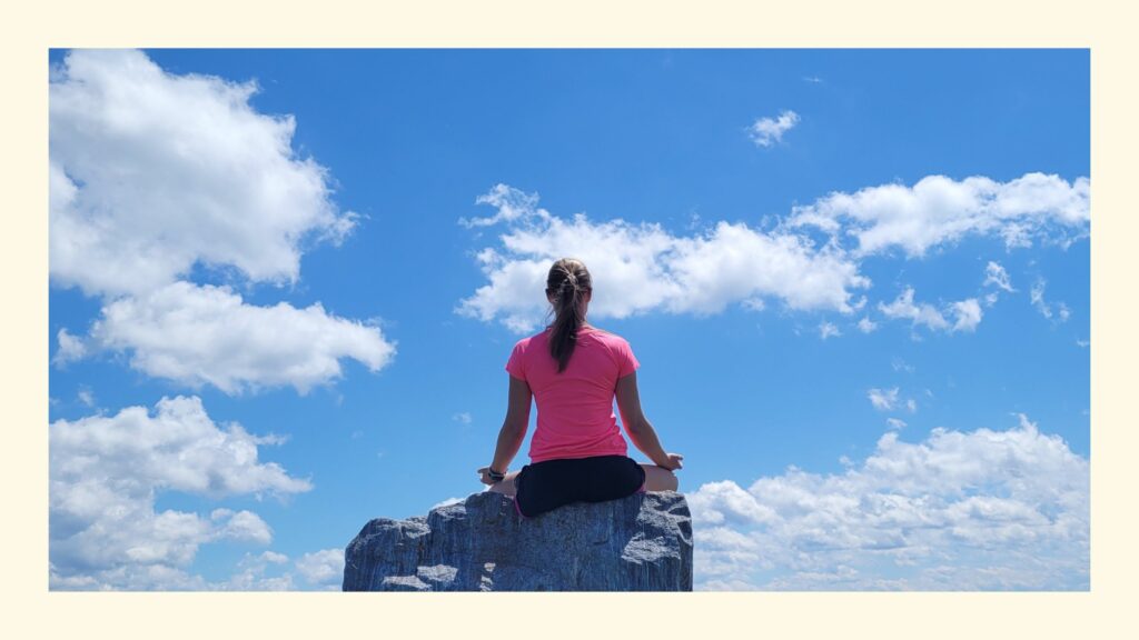 Rückansicht einer Frau im Sitzen auf Fels bei einer Meditation für Anfänger, über ihr blauer Himmel mit weißen Wolken