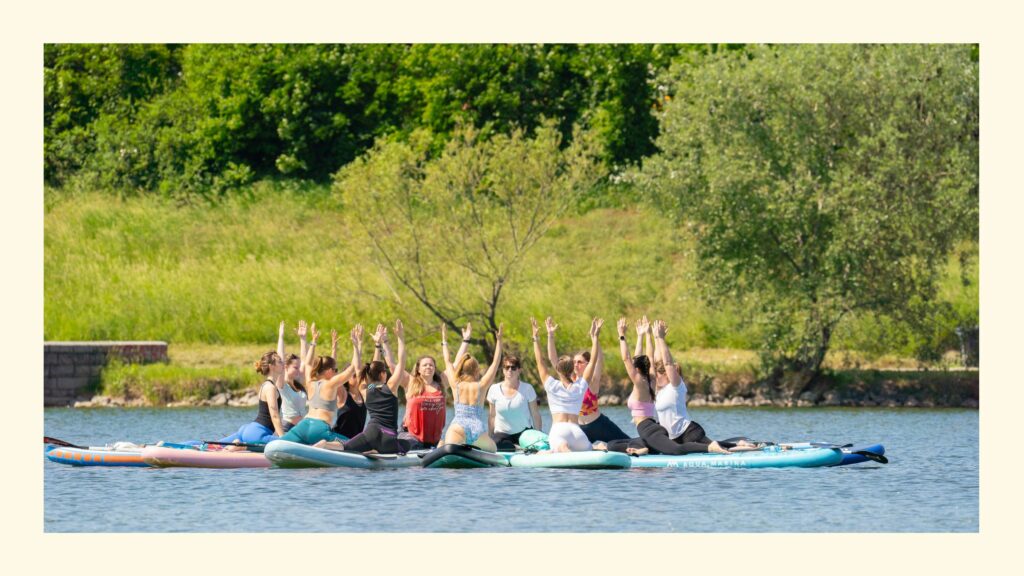 Gruppe von Frauen auf sternförmig verbundenen SUP Boards am Wasser übt die Yoga Taube mit nach oben gestreckten Armen, im Hintergrund Bäume und Wiese