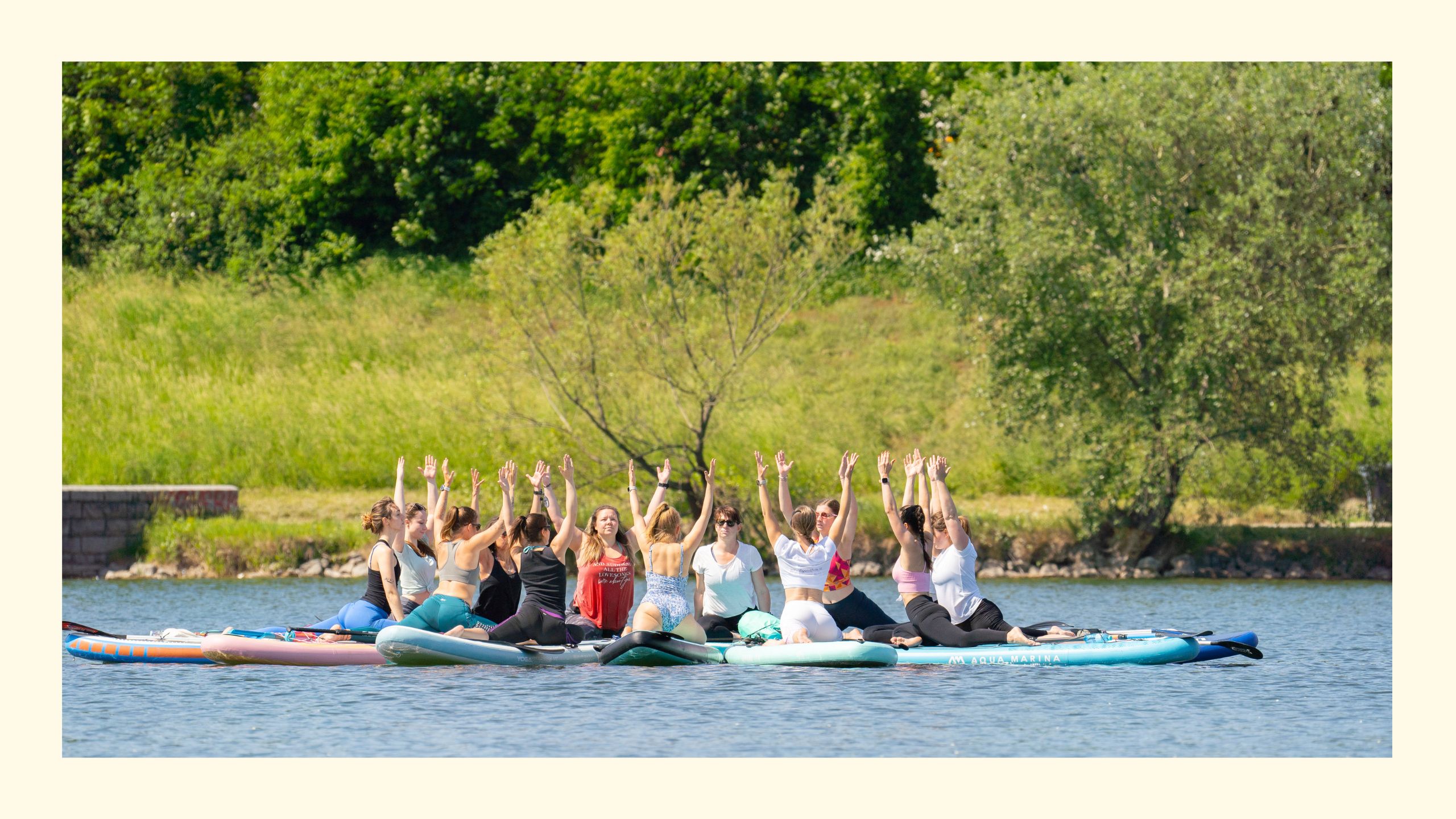 Gruppe von Frauen auf sternförmig verbundenen SUP Boards am Wasser übt die Yoga Taube mit nach oben gestreckten Armen, im Hintergrund Bäume und Wiese