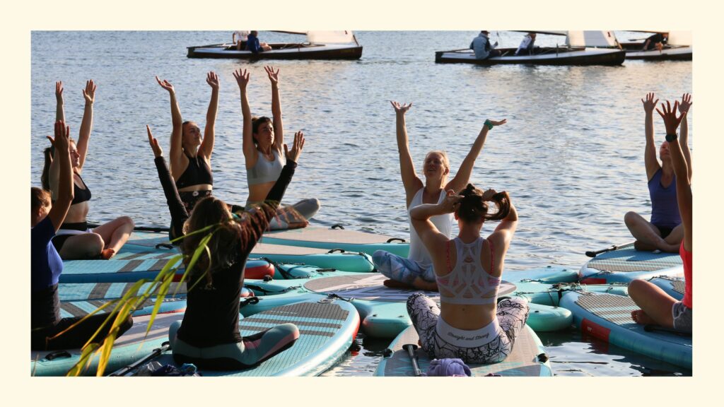 Sabrina mit ihren SUP Yoga Schülerinnen auf den Boards sitzend mit ausgestreckten Armen in Wien 2025, im Hintergrund Segelboote