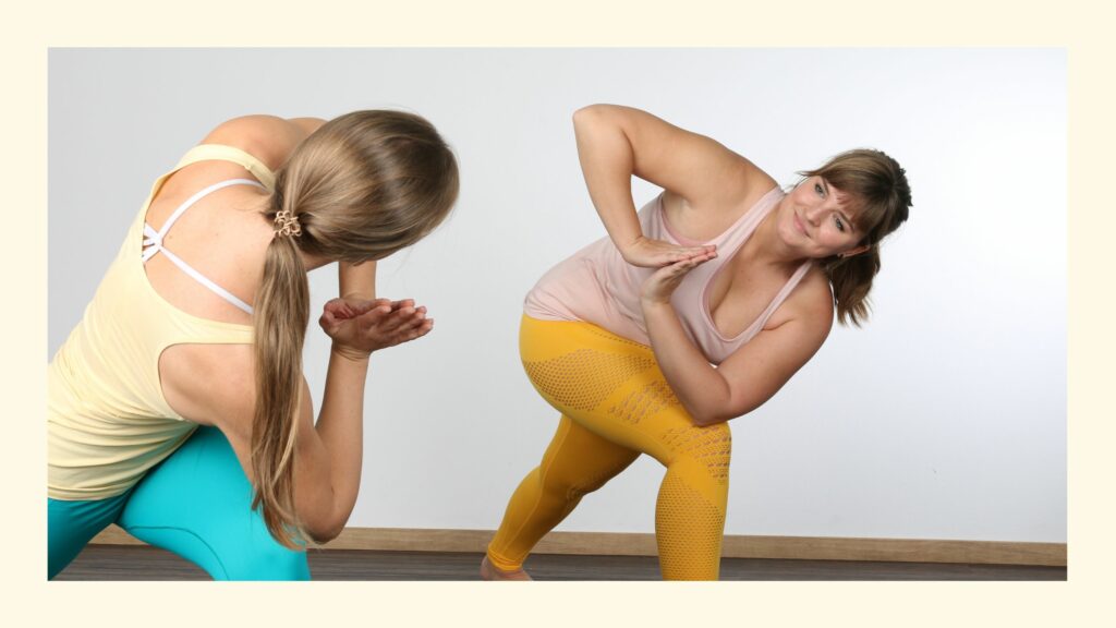 Ausbildnerin Sabrina und Teilnehmerin lächeln einander in gedrehtem Ausfallschritt an bei der Yogalehrer Ausbildung in Österreich und online
